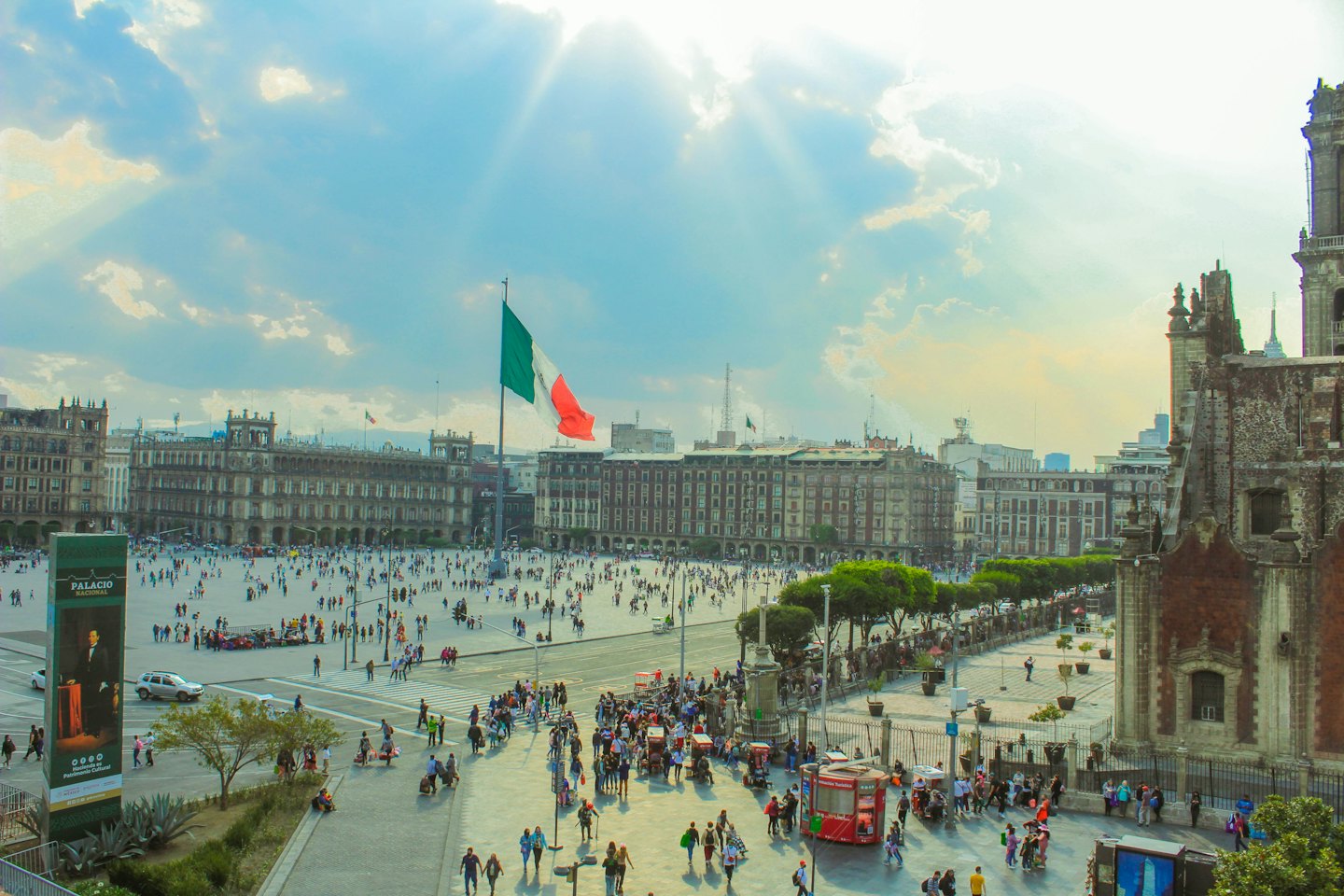 Bandera mexicana ondeando en una plaza urbana concurrida con edificios de piedra.